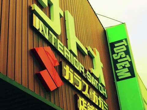Raised pale interior shop lettering and a red emblem decorate brown siding beside a tall green sign.