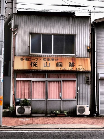 A narrow corrugated shop has a faded tan sign above pink curtains and two ground-level air conditioners.