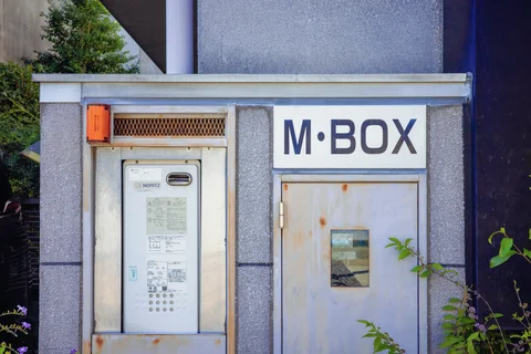 A gray metal enclosure has an intercom panel, orange light, and a rust-streaked door beneath bold lettering.
