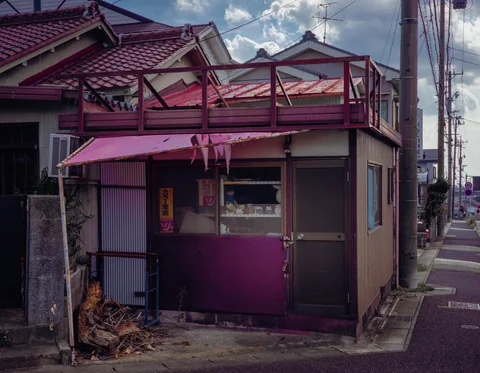 A small weathered corner building has a red rooftop railing and a faded pink awning.