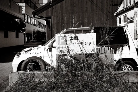 A deteriorating white car sits behind weeds with a parking sign against its side in black and white.