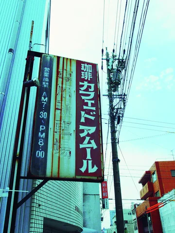 A rust-streaked vertical cafe sign projects beside blue siding beneath dense overhead utility wires.