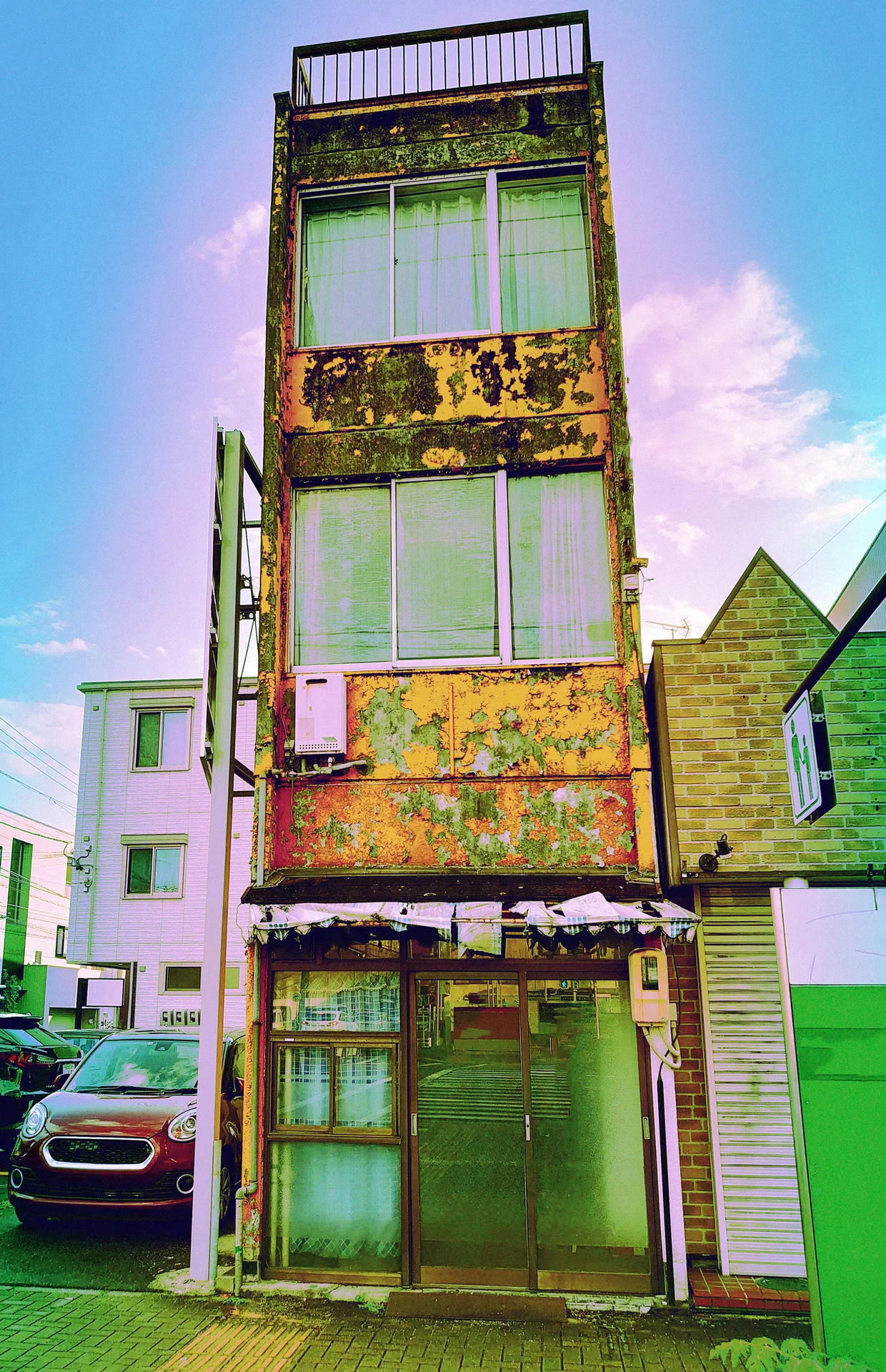 A very narrow multi-story building has peeling yellow paint, green windows, and a dark rooftop railing.