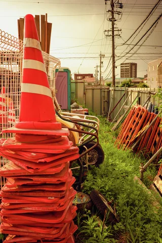 Orange traffic cones are stacked beside metal barriers and weeds in a fenced storage area.