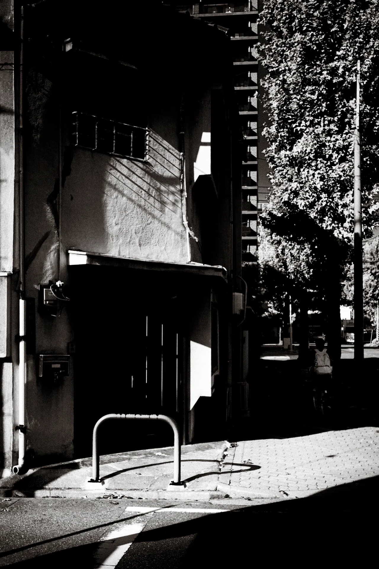 Sunlight crosses a narrow sidewalk and metal barrier beside a deeply shadowed building entrance.