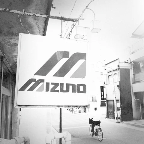 A large Mizuno sign projects over a narrow street where a cyclist passes in black and white.