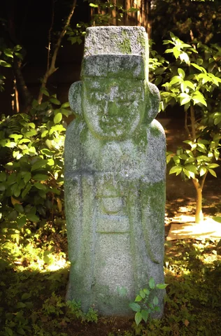A moss-stained stone figure with a square headdress stands amid sunlit leaves and shaded shrubs.