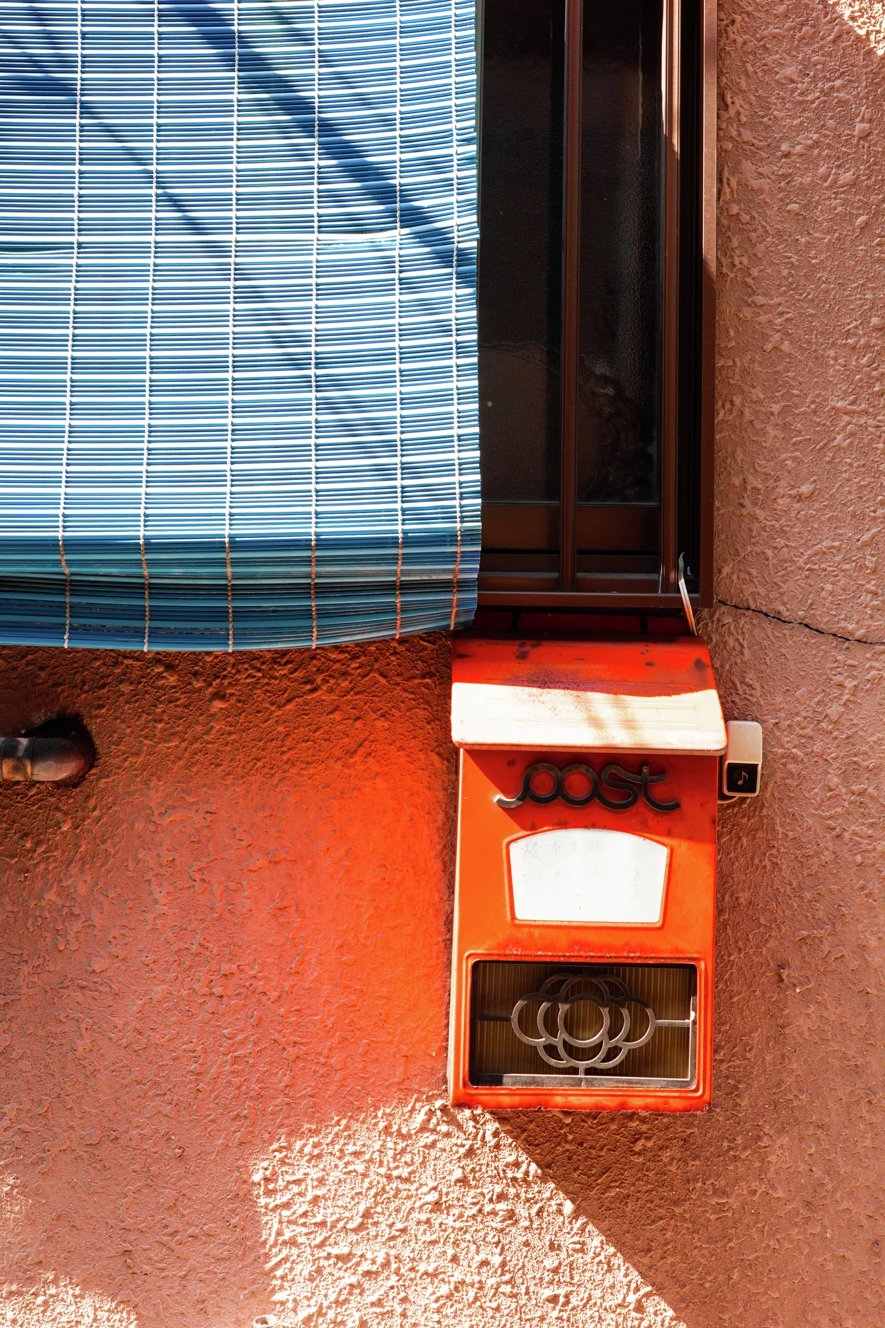 A bright red mailbox casts a diagonal shadow on an orange wall beneath a blue window shade.