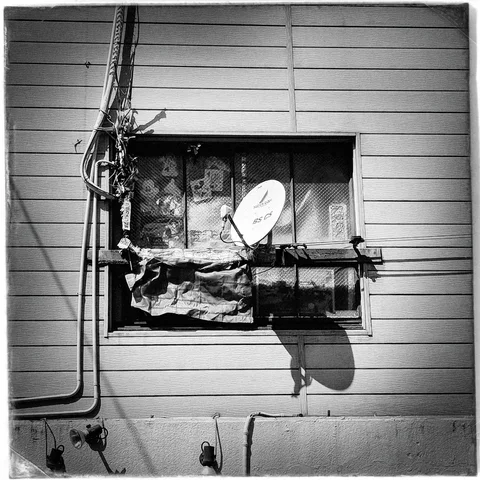 A satellite dish and tangled cables cluster around a patched window on horizontal siding in black and white.