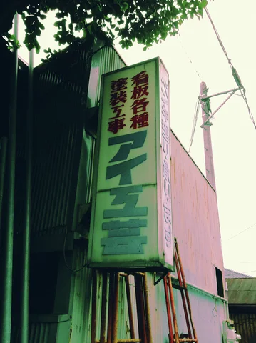A tall faded sign bearing colored Japanese lettering projects beside corrugated siding beneath leafy branches.
