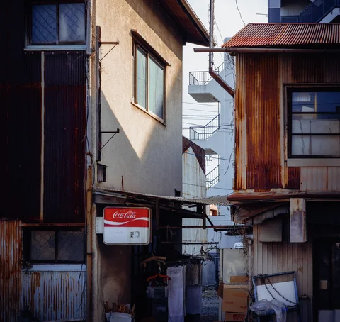 A narrow passage separates weathered buildings with corrugated siding, small windows and a red drinks sign.