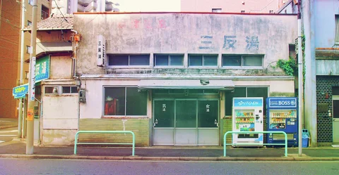 Two vending machines stand beside a low weathered building with green doors and pale turquoise roadside railings.