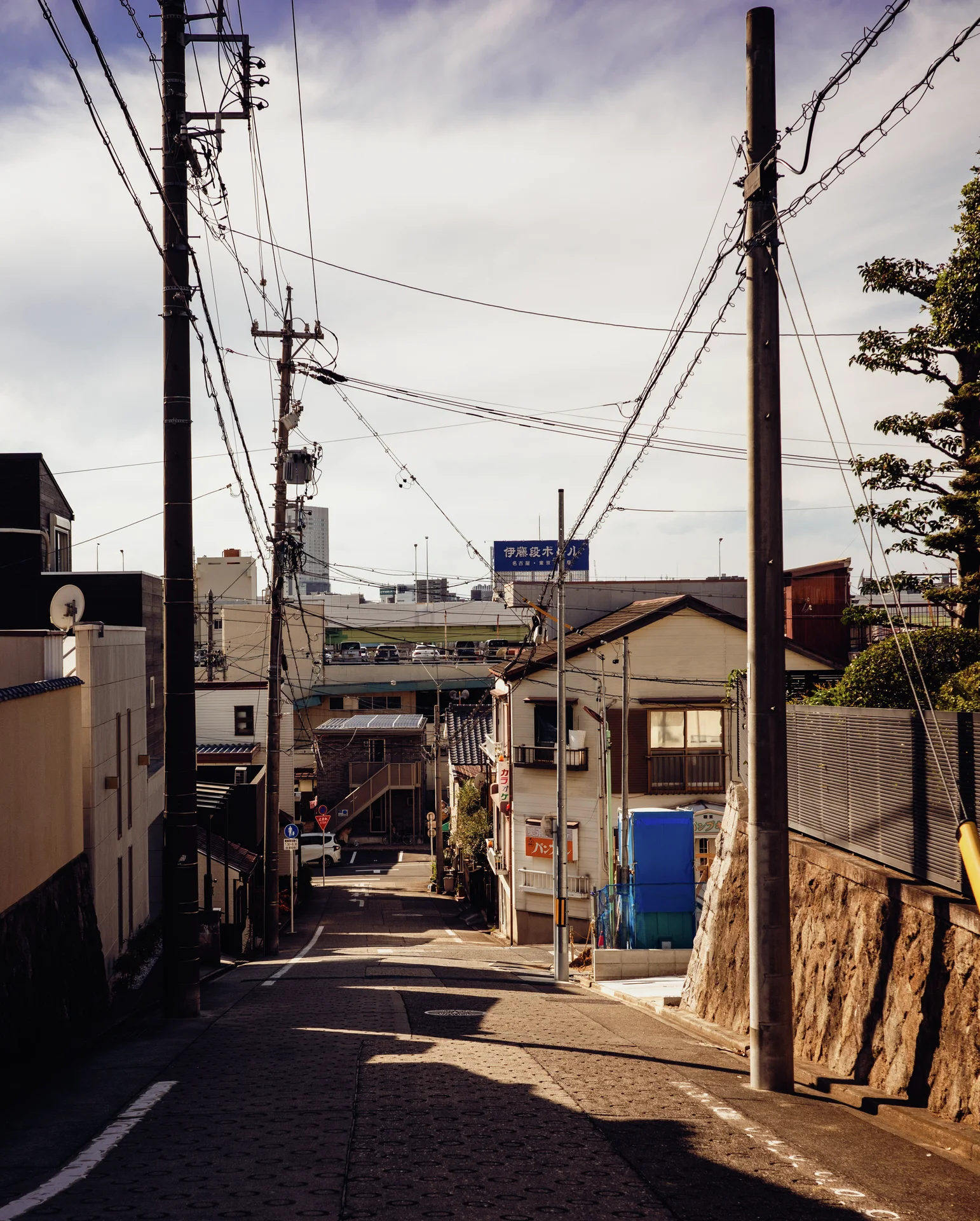 A narrow sloping street passes small buildings beneath utility poles and tangled overhead cables.