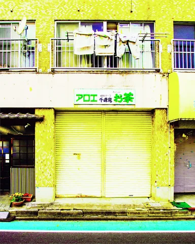 Laundry hangs from an upper balcony above closed yellow shutters and a white sign with green lettering.