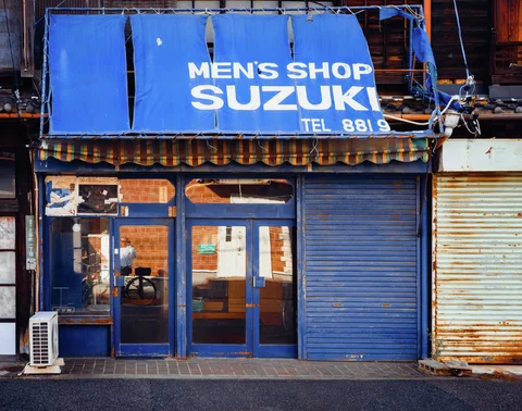A blue shopfront has a torn fabric awning above glass doors and a closed metal shutter.