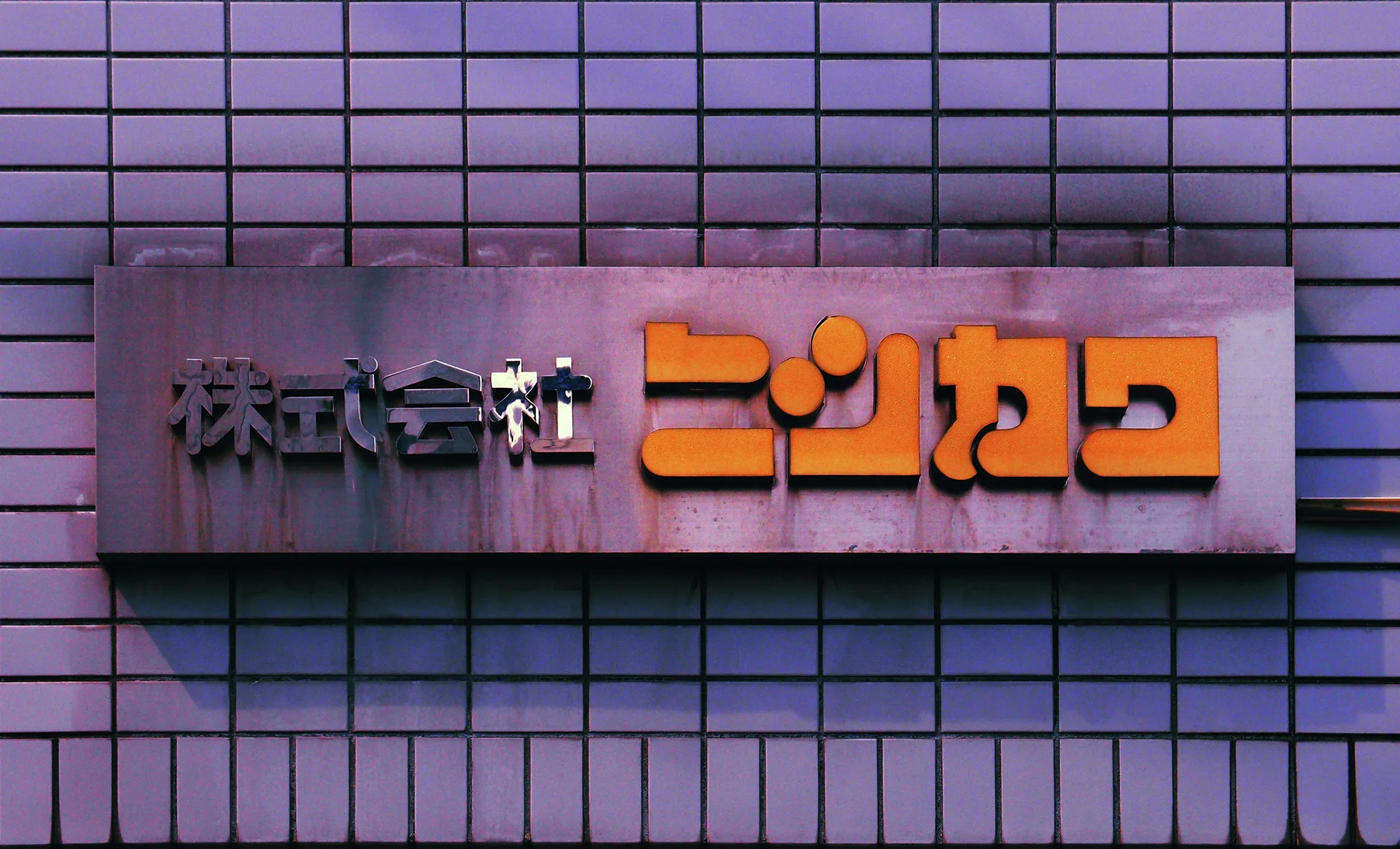 Raised orange Japanese lettering and smaller dark characters sit on a gray plaque against square wall tiles.