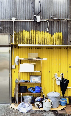 Buckets, containers, and umbrellas cluster beneath pipes on a yellow corrugated exterior wall.