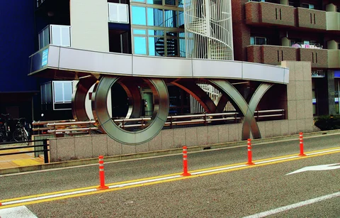 Large curved metal sculptures stand beneath a modern entrance canopy beside a road lined with orange bollards.