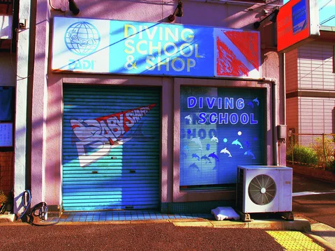 Blue diving-school signs and illustrated shutters mark a small shop beside an outdoor air conditioner.