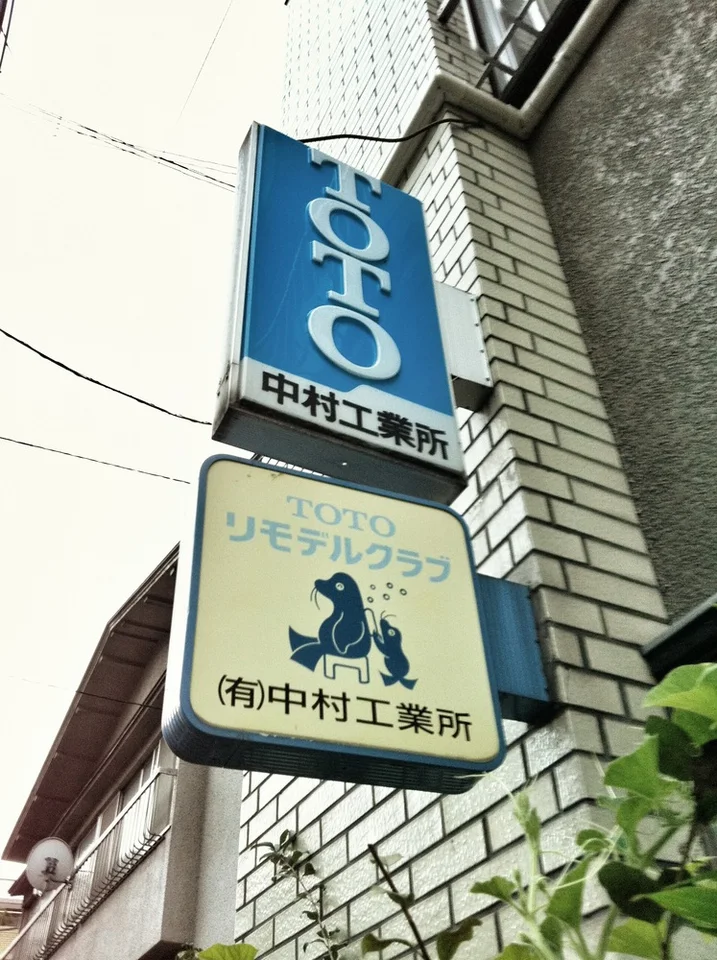 Two projecting signs hang beside a pale brick wall, including a blue vertical sign and illustrated lower panel.