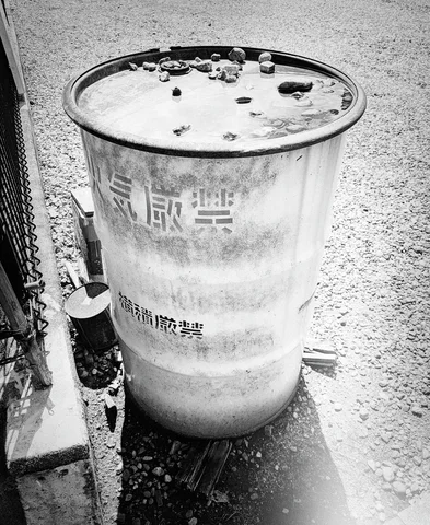 A weathered metal drum holds water and floating debris beside a roadside grate in black and white.