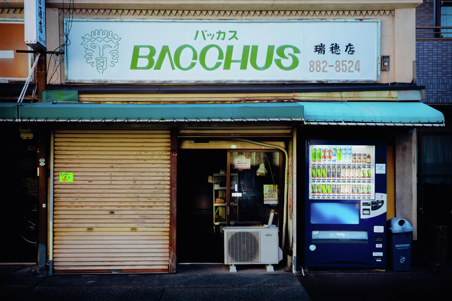 A BACCHUS sign tops a small shop with closed shutters and a blue vending machine.