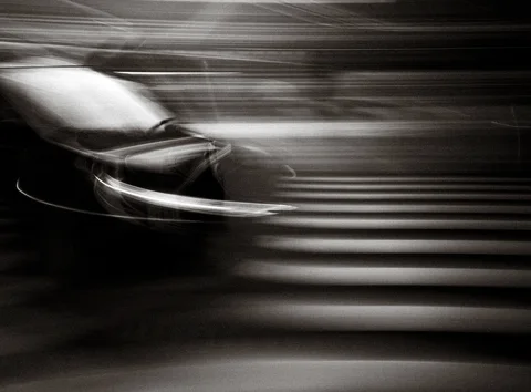 A dark car blurs across broad striped road markings in a streaked black and white street scene.