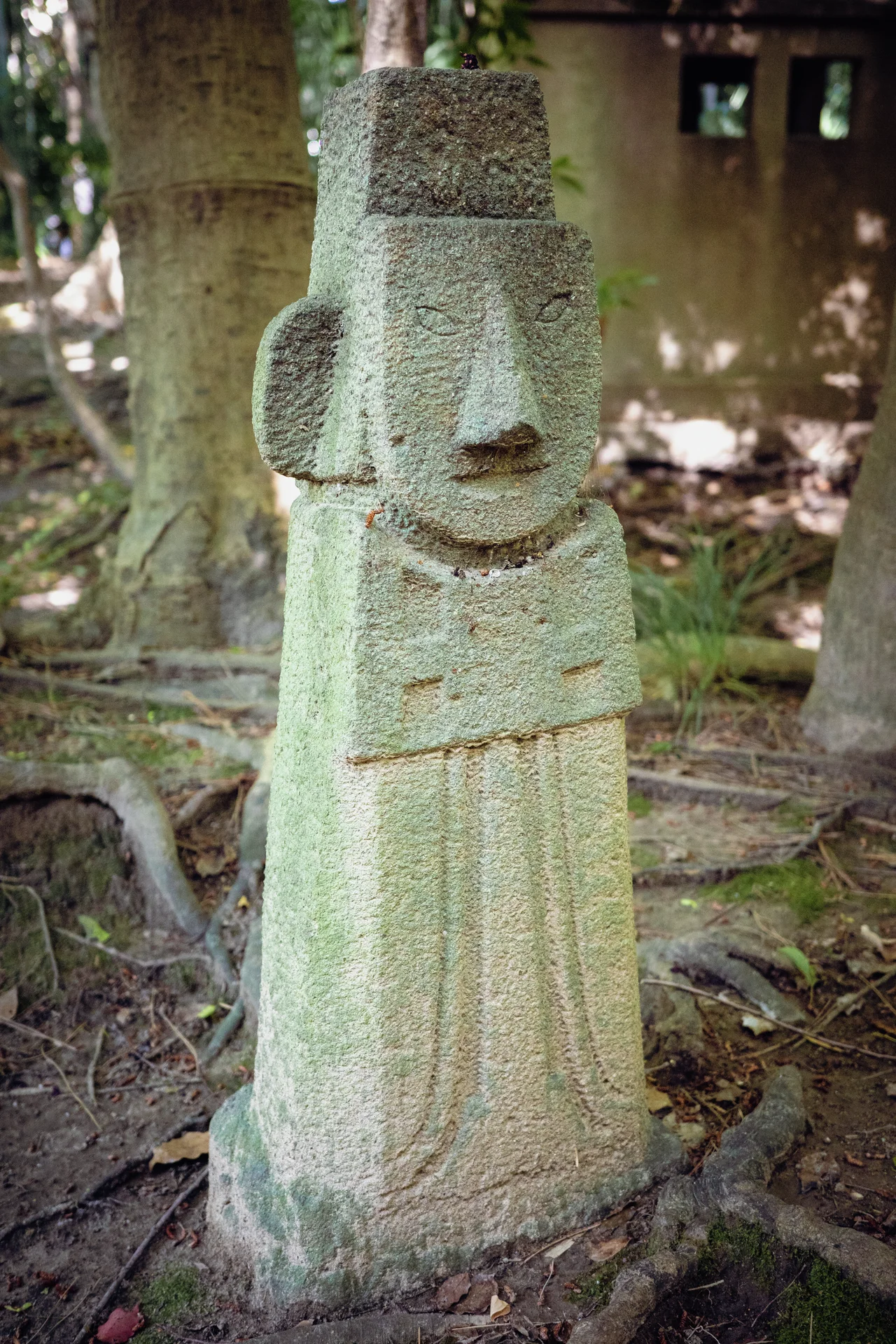 A pale carved stone figure with elongated ears stands on a base beside exposed tree roots.