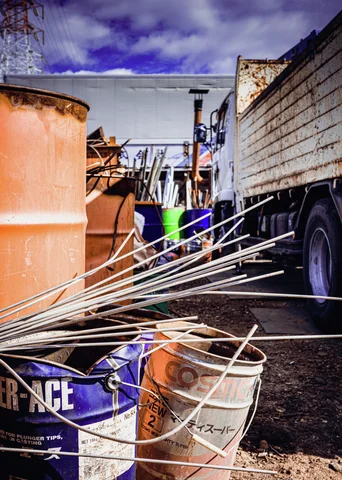 Long metal rods cross buckets beside an orange tank and a truck in a cluttered outdoor work area.