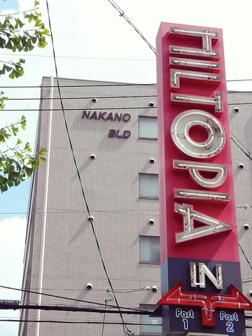 A tall pink sign with raised pale vertical letters rises beside a gray building and overhead wires.
