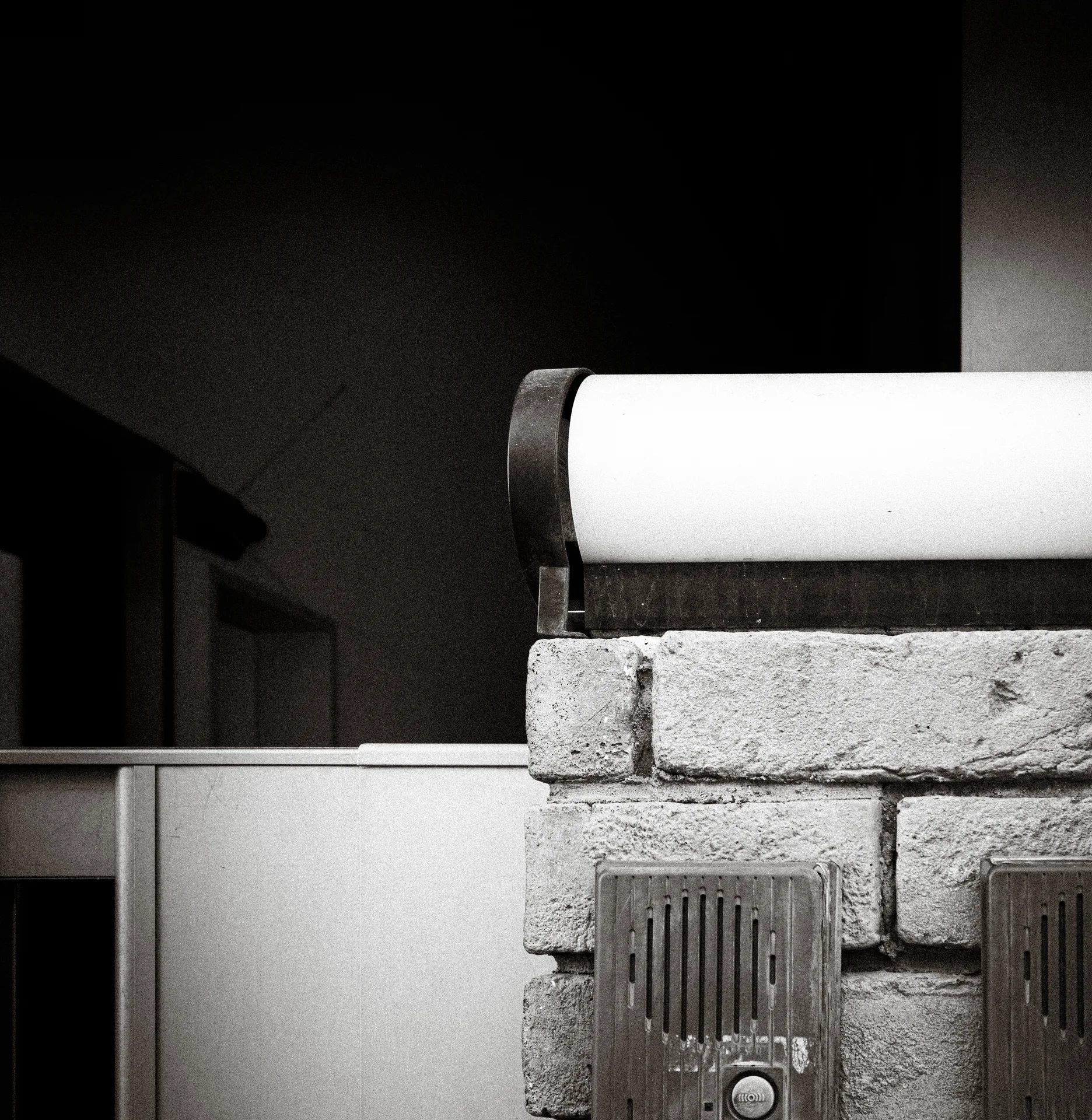 A pale curved cover rests above a rough block wall and metal vent in black and white.