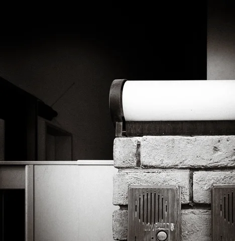 A pale curved cover rests above a rough block wall and metal vent in black and white.