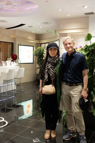 Two adults pose beside indoor greenery, with glossy dark floor tiles reflecting lights and nearby white furniture.