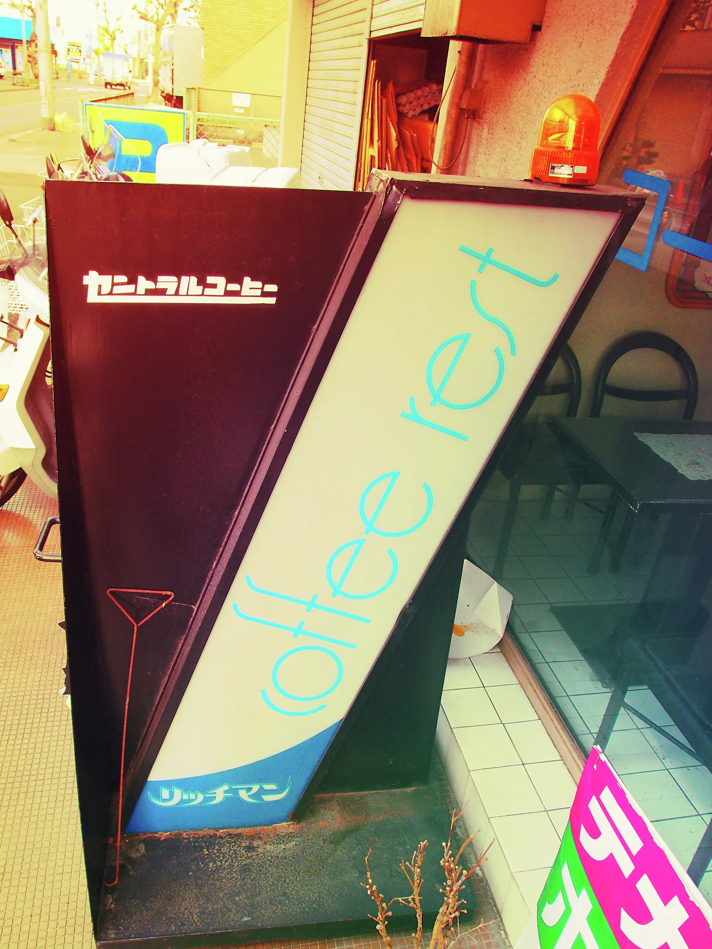 A tall angled cafe sign with turquoise lettering stands beside glass and an orange light.