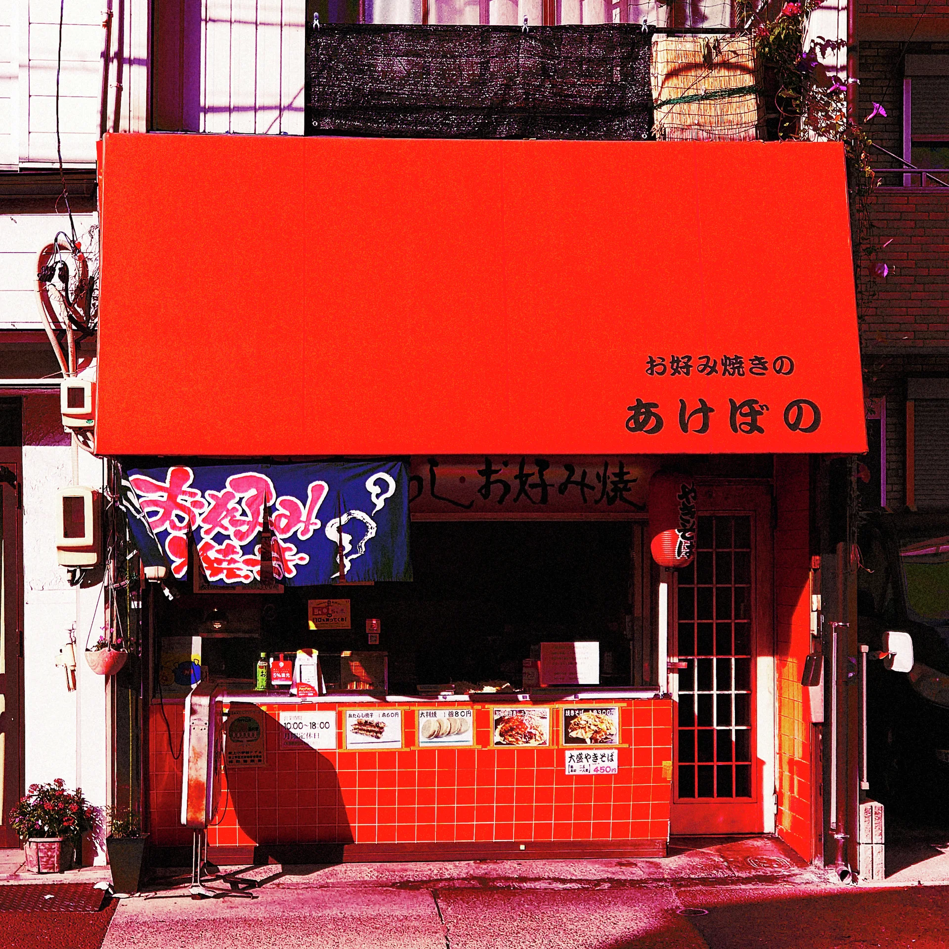A broad red awning shelters a small open food counter faced with red tiles and illustrated signage.