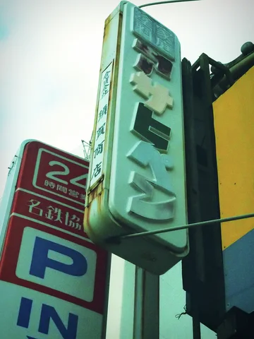 A weathered turquoise vertical sign with raised Japanese lettering hangs beside a red parking entrance sign.