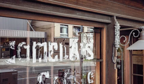 Distressed white lettering covers a wooden-framed shop window reflecting bicycles and buildings across the street.