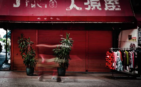 A blurred passerby crosses closed red shutters between potted plants and a rack of clothing.