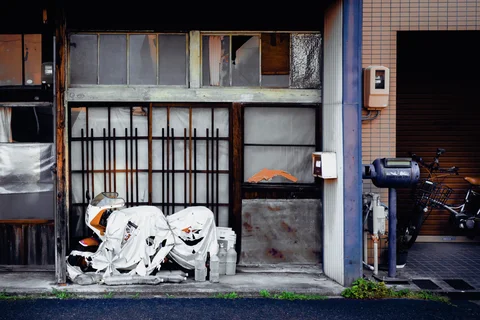 A white-covered scooter sits outside weathered sliding panels beside a narrow tiled entrance and bicycles.