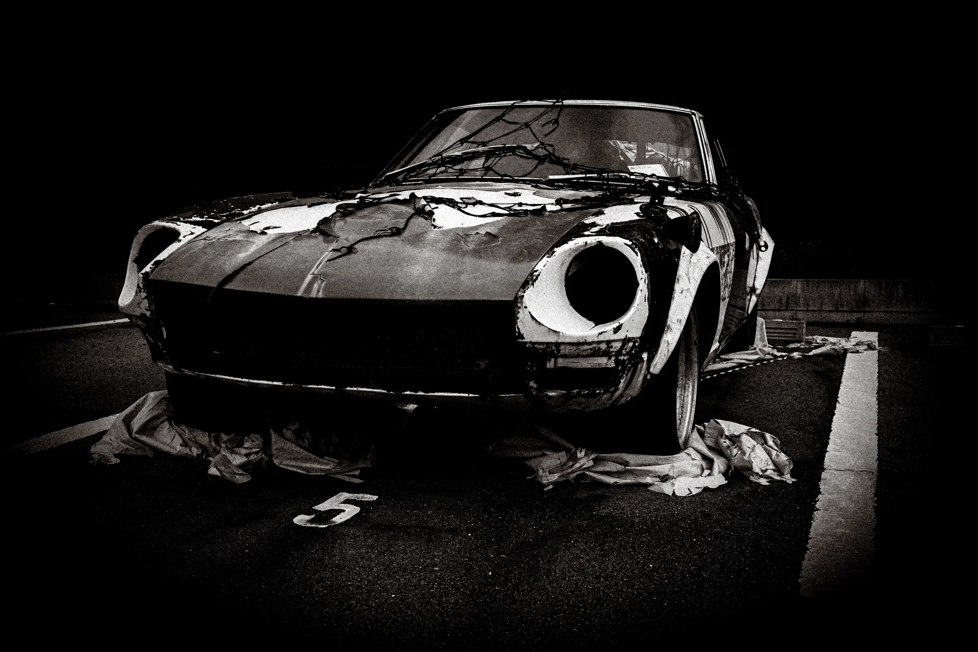 A deteriorated sports car with empty headlight openings sits amid loose coverings in black and white.