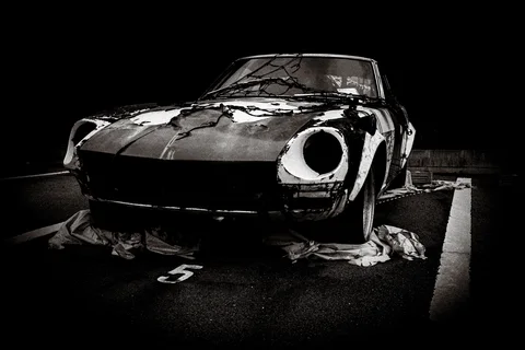 A deteriorated sports car with empty headlight openings sits amid loose coverings in black and white.
