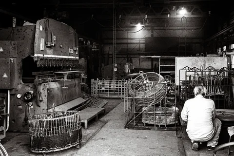 A crouching person faces stacked wire frames beside large machinery inside a workshop in black and white.