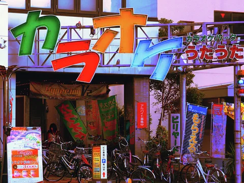 Large multicolored Japanese characters span a karaoke entrance crowded with bicycles, banners and small signs.
