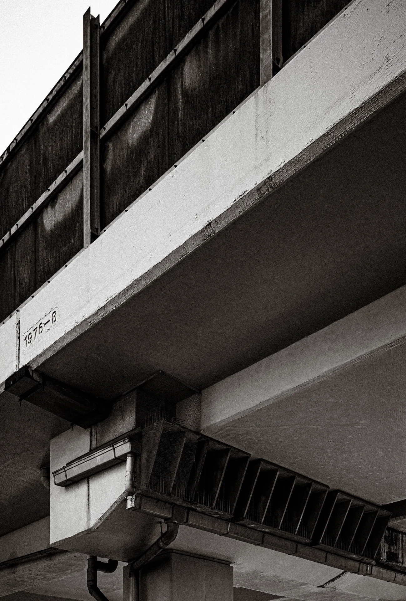 Angular concrete ledges project above a metal cable tray on a building facade in black and white.