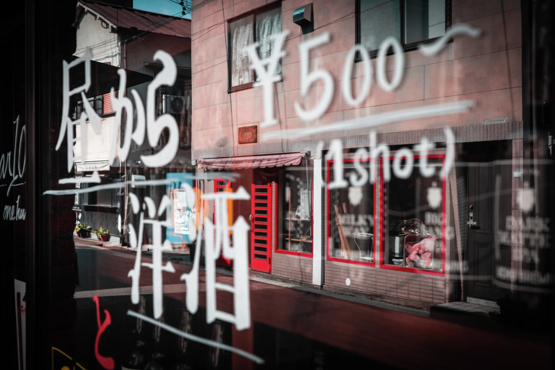 White handwritten Japanese lettering on glass overlaps a reflected building with red window frames and an awning.