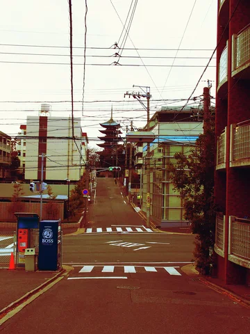 A narrow street with white crossing stripes leads toward a distant tiered tower beneath overhead wires.