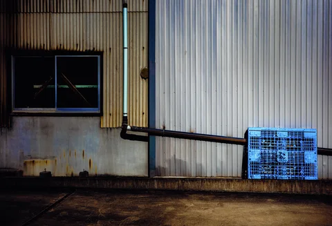 A blue plastic crate rests against corrugated siding crossed by a black drainpipe beside a small window.