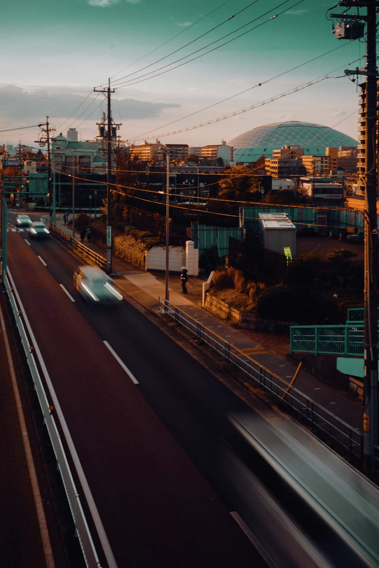 Blurred white vehicles streak along a road beside low buildings and utility poles beneath a pale green sky.