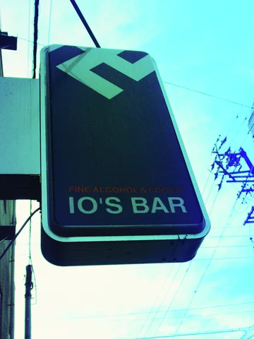 A dark projecting bar sign with a pale geometric logo hangs against a turquoise sky and wires.