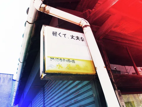 A faded white and yellow sign hangs beneath rusty pipes above a blue shutter.